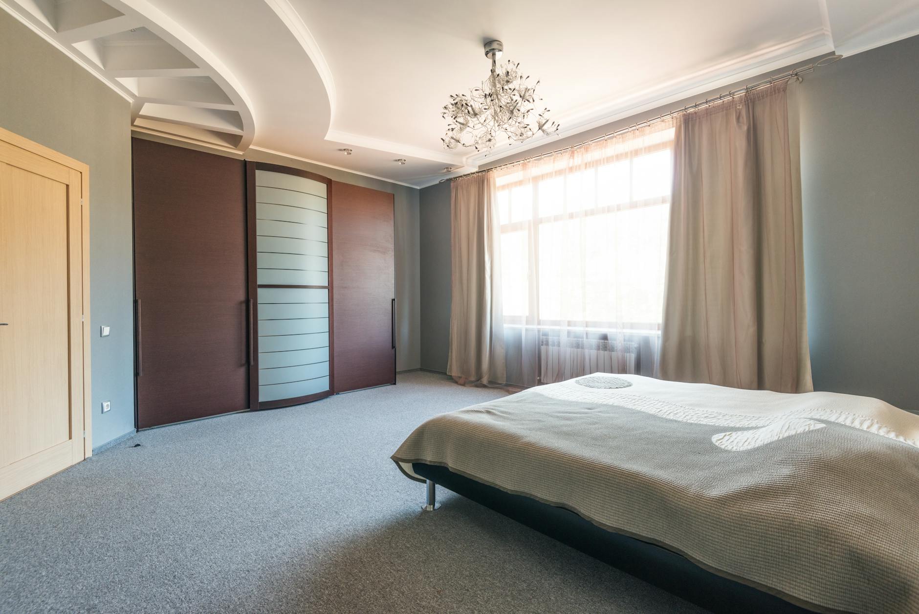 Spacious modern bedroom with platform bed, large curtained window, decorative chandelier and sliding wood wardrobe